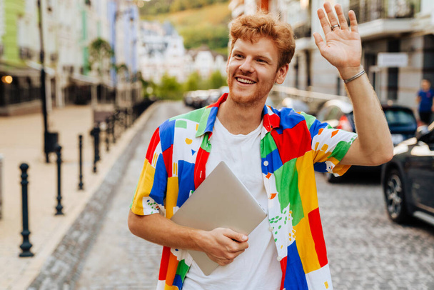 Portrait of young handsome smiling redhead man holding laptop, waving and looking aside, while standing on the street - Фото, изображение
