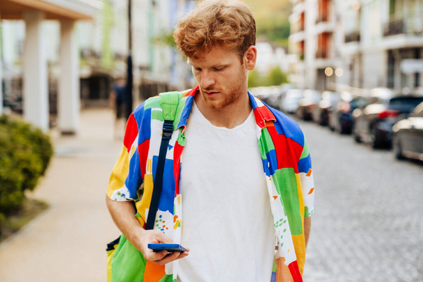 Portrait of young handsome calm redhead man in colofrul shirt holding and using his phone, while stending on the street - Fotografie, Obrázek