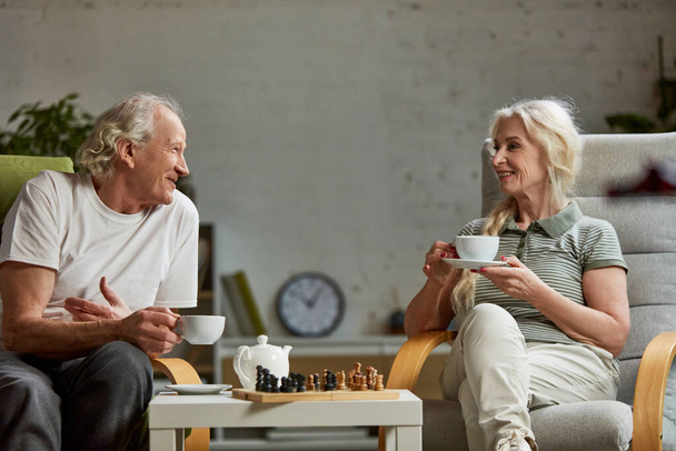 Happy elderly, senior couple, retired man and woman spending time at home, playing chess and talking together. Leisure time and hobby. Concept of family, relationship, retirement, lifestyle, happiness - Zdjęcie, obraz