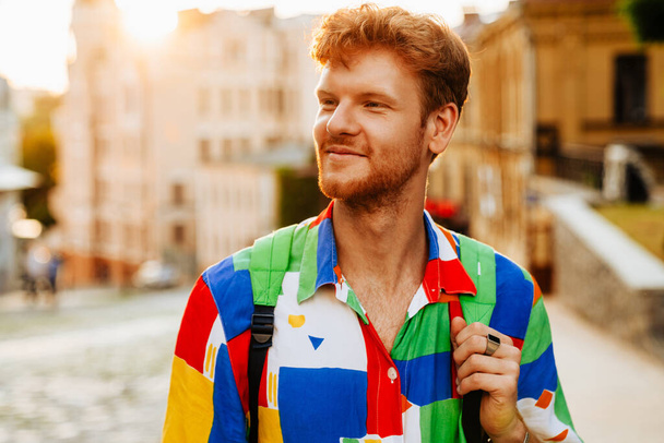Portrait of young handsome calm redhead man in colorful shirt looking aside, while standing in the sun on the street - 写真・画像