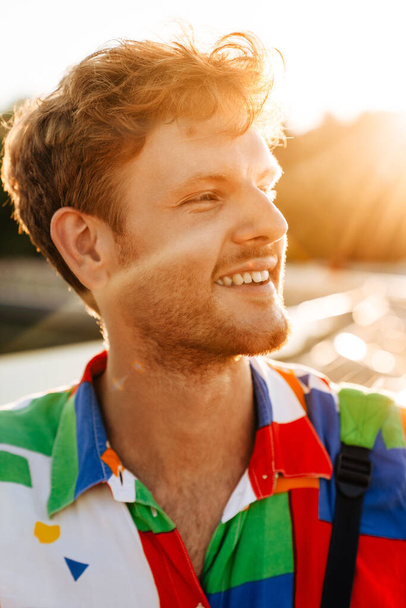 Portrét mladého pohledného usmívajícího se šťastného muže v pestré košili, dívajícího se stranou, zatímco stojí na slunci - Fotografie, Obrázek