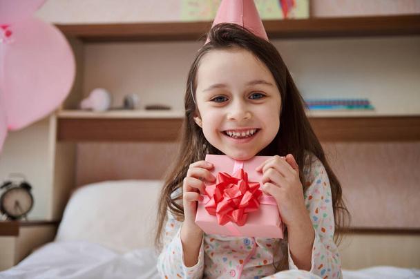 Menina adorável criança em pijama e chapéu festivo rosa, gentilmente abraçando seu presente de aniversário, sorrindo amplamente olhando para a câmera, sentada na cama em seu quarto com balões de ar infláveis - Foto, Imagem