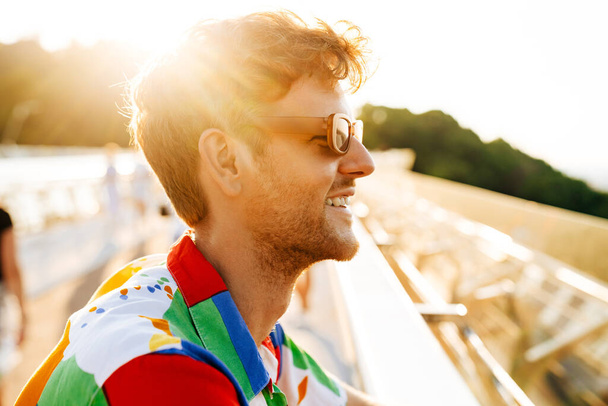 Profile portrait of handsome smiling man in sun glasses and colorful shirt sitting in the sun - Zdjęcie, obraz