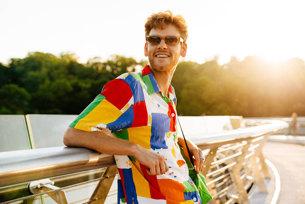 Profil portretu przystojnego uśmiechniętego mężczyzny w okularach przeciwsłonecznych i barwnej koszuli opierającego się na balustradzie i patrzącego na bok, stojącego w słońcu - Zdjęcie, obraz