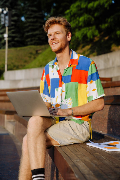 Jeune homme élégant et souriant avec ordinateur portable regardant de côté, assis avec les jambes croisées sur tribune dans le parc - Photo, image