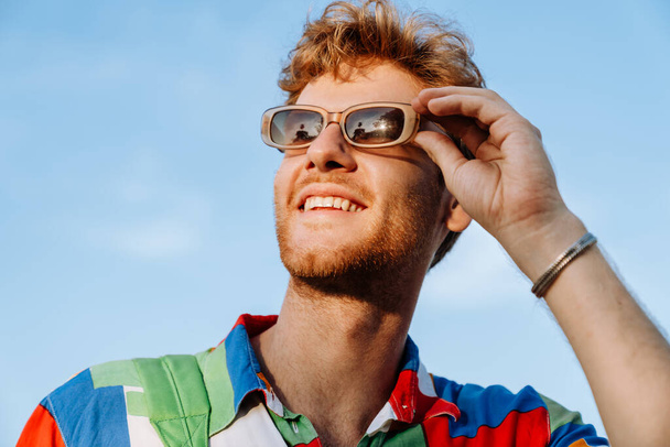 Portrait of young handsome stylish smiling man in colorful shirt adjusting his sun glasses and looking aside - 写真・画像