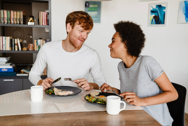 Young beautiful smiling interracial couple looking at each other during breakfast. Young handsome redhead guy and beautiful african curly woman at home - 写真・画像