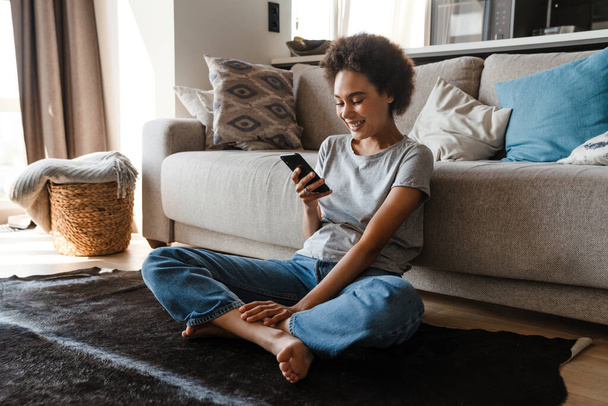 Young beautiful smiling curly african woman holding and using her phone sitting on carpet on the floor in lotus pose leaning on couch in cozy living room at home - Φωτογραφία, εικόνα