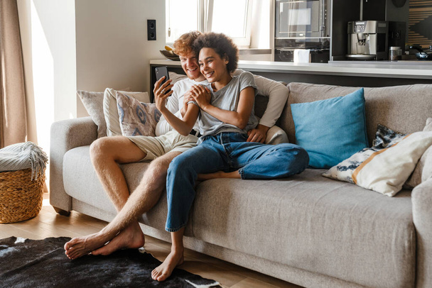 Young happy interracial couple watching video on phone together, while sitting on sofa in cozy living room at home - Foto, Imagem