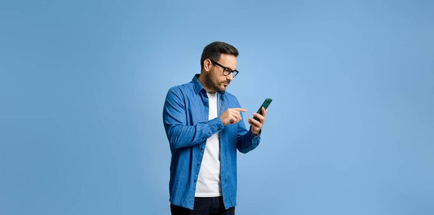 Серьезный красивый молодой человек, одетый в джинсовую рубашку серфинг интернет через смартфон, находясь в изоляции на синем фоне - Фото, изображение