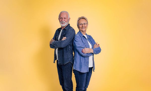 Smiling senior couple dressed in denim shirts with arms crossed standing and posing confidently while standing back to back isolated over yellow background - Photo, Image