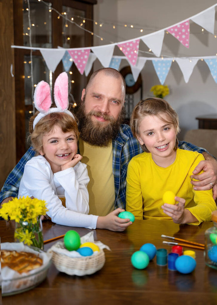 Easter Family traditions. Father and two caucasian happy children with bunny ears dye and decorate eggs with paints for holidays while sitting together at home table. Kids embrace and smile in cozy. - Φωτογραφία, εικόνα
