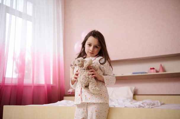 Rozkošné kavkazské dítě, krásná holčička ve stylovém bílém pyžamu s barevnými tečkami, objímající své plyšové ovečky a roztomile se usmívající, dívající se do kamery, stojící ve svém útulném světlém pokoji interiéru - Fotografie, Obrázek