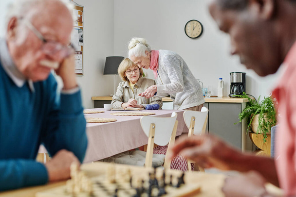 Yaşlı kadınlar örgü örmeyi tartışırken yaşlı erkekler huzurevinde satranç oynuyorlar. - Fotoğraf, Görsel
