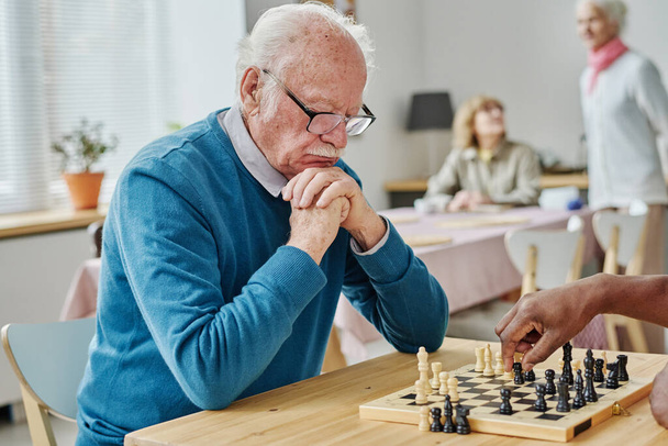 Σκεπτικός ηλικιωμένος με γυαλιά, συγκεντρωμένος στο σκάκι, παίζει με τον φίλο του στο τραπέζι. - Φωτογραφία, εικόνα