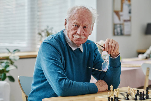 Retrato de hombre mayor con el pelo gris mirando a la cámara mientras juega al ajedrez en la mesa - Foto, Imagen