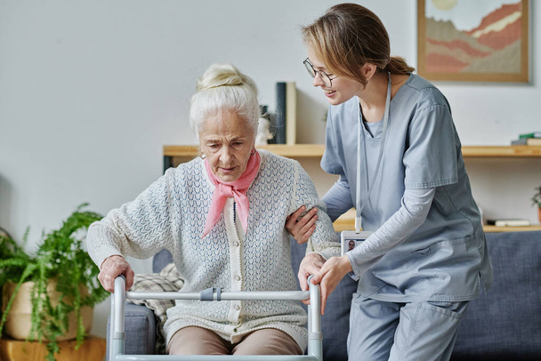 Молодая волонтёр, работающая в доме престарелых, помогает пожилой женщине встать с помощью ходунков - Фото, изображение