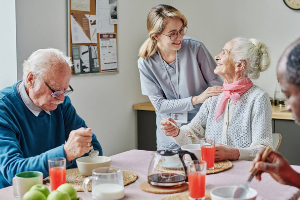 Cuidador hablando con personas mayores y ayudándoles mientras comen en la mesa - Foto, imagen