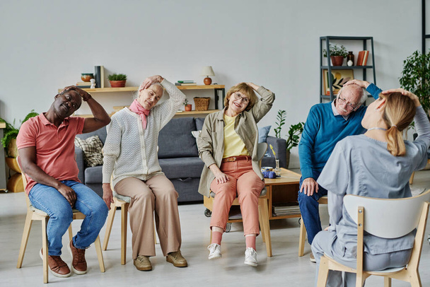Ryhmä vanhempia ihmisiä istuu tuoleilla ja tekee liikuntaa yhdessä ohjaajan kanssa - Valokuva, kuva