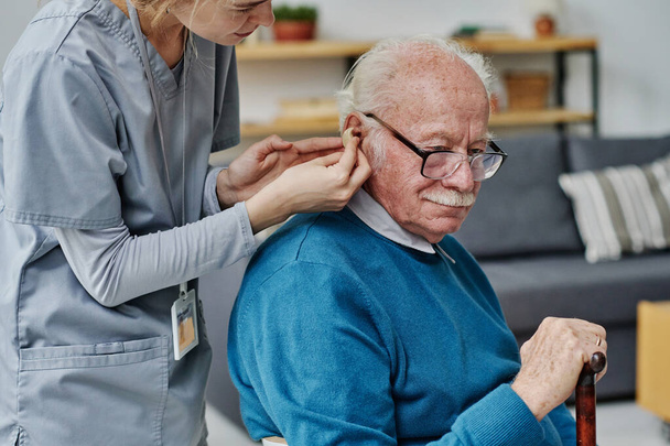 Young caregiver helping to wear hearing aid to senior man while she caring about senior people in nursing home - Фото, зображення