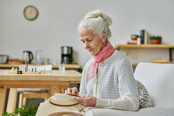 Ηλικιωμένη γυναίκα συγκεντρωμένη στη χειροτεχνία της, κάθεται στην πολυθρόνα και κεντάει - Φωτογραφία, εικόνα