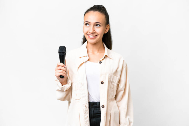 Joven cantante mujer recogiendo un micrófono sobre aislado fondo blanco pensando una idea mientras mira hacia arriba - Foto, Imagen
