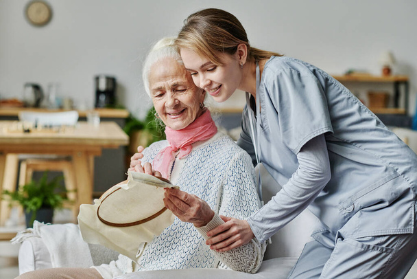 Χαμογελαστή ηλικιωμένη γυναίκα κεντάει και δείχνει τη χειροτεχνία της στον φροντιστή - Φωτογραφία, εικόνα