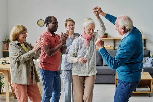 Старшая пара танцует вместе, в то время как другие люди хлопают в ладоши во время отдыха в доме престарелых - Фото, изображение