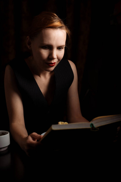 A woman sits at a table with a book and tea and cake in the dark. A woman of forty years old relaxes alone. - Fotó, kép