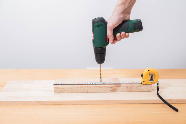 Un hombre trabaja con un destornillador eléctrico en una mesa de madera, también hace mediciones con una cinta métrica - Foto, Imagen