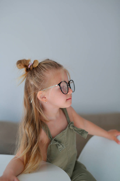 une belle fille élégante de trois ans, portant des lunettes, assise à une table blanche, pense, regarde l'appareil photo, pose pour un photographe - Photo, image
