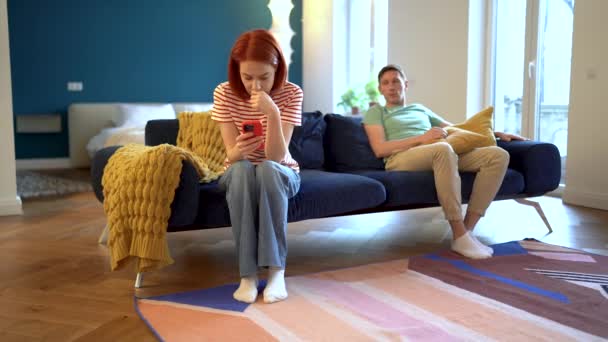 Offended woman after quarrel with husband sits stooped on couch, ignoring looks at screen smartphone smiling man wants to make peace flirting throwing sofa cushion at upset wife attracting attention - Footage, Video