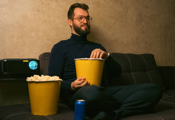 Een knappe man met een bril die een moderne videoprojector gebruikt, een film kijkt, een hapje eet. Een man met een baard die popcorn eet en films kijkt. bioscoop - Foto, afbeelding