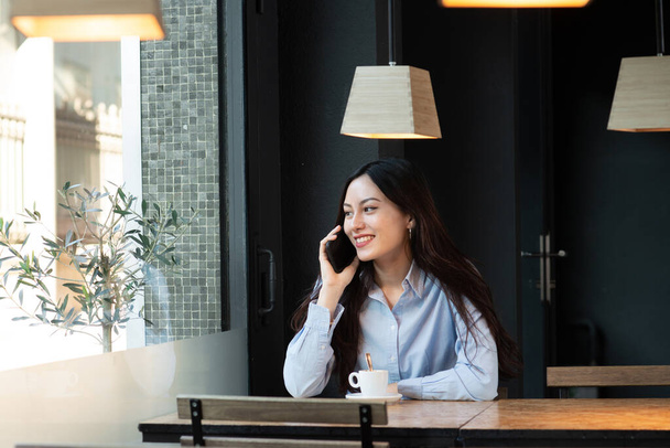 Asiática joven habla por teléfono mientras bebe café en la cafetería - Foto, imagen