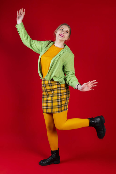 Симпатичная красивая девушка в зеленой и желтой одежде позирует на изолированном красном фоне. Счастливая улыбка, одна нога вверх, руки вверх. - Фото, изображение