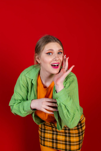 A cute beautiful girl in a green and yellow clothes posing against an isolated red background. Whispering. gossiping. telling a secret. - Fotó, kép
