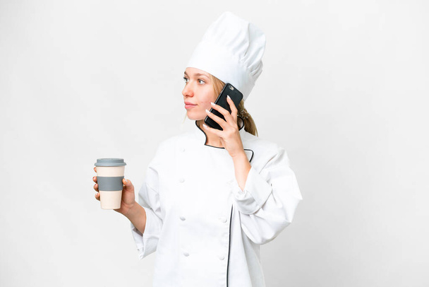 Молодая женщина шеф-повар на изолированном белом фоне держа кофе, чтобы забрать и мобильный - Фото, изображение