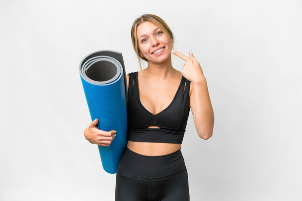 Blonde sport woman going to yoga classes while holding a mat over isolated white background giving a thumbs up gesture - Foto, imagen