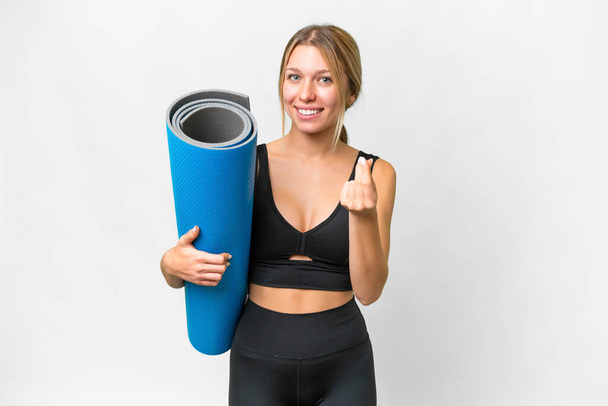 Blonde sport woman going to yoga classes while holding a mat over isolated white background making money gesture - Foto, afbeelding