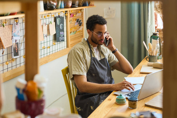 Young male clerk in apron calling clients and looking through online orders while sitting by workplace in front of laptop - 写真・画像