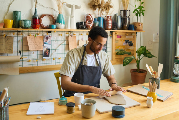 Młody wieloetniczny mężczyzna w odzieży roboczej spłaszczającej gliny do tworzenia nowych ręcznie robionych przedmiotów ceramicznych podczas siedzenia w miejscu pracy - Zdjęcie, obraz