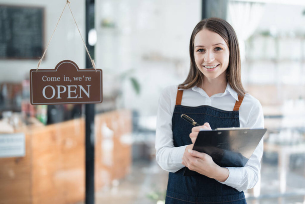 Ξεκινώντας και ανοίγοντας μια μικρή επιχείρηση, μια νεαρή Ασιάτισσα δείχνει ένα χαμογελαστό πρόσωπο σε μια ποδιά που στέκεται μπροστά από έναν πάγκο καφετέριας. Ιδιοκτήτης επιχείρησης, Εστιατόριο, Barista, Cafe, Online ΜΜΕ. - Φωτογραφία, εικόνα