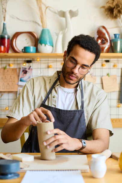 Creatieve mannelijke kruik maken van nieuwe klei vaas of kan tijdens het zitten aan tafel met benodigdheden voor creatibe aardewerk in studio of werkplaats - Foto, afbeelding