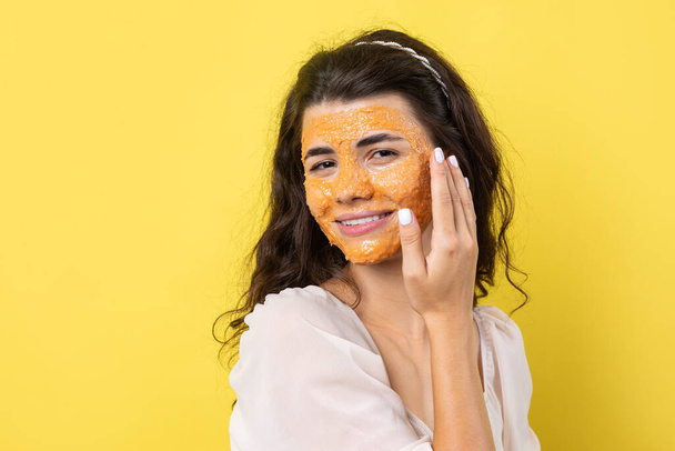 Een jong brunette meisje met een reinigende scrub masker op haar gezicht, kijkt naar de camera en glimlacht, tegen een gele achtergrond. - Foto, afbeelding