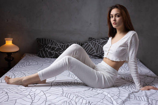 Uma jovem garota atraente vestida com pijama branco está descansando em uma cama em um quarto na iluminação da noite. - Foto, Imagem