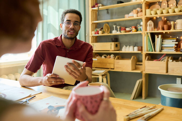 Молодой черный мужчина с планшетом смотрит на коллегу-женщину во время обсуждения идей для нового творческого проекта в мастерской - Фото, изображение