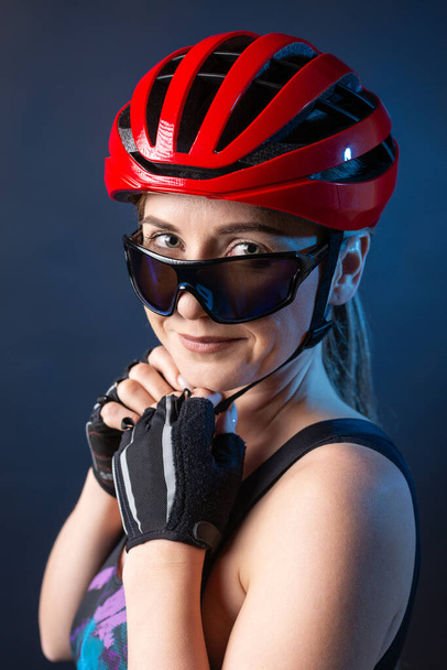 A young female cyclist wearing a safety helmet and glasses, dressed in a bib shorts poses against a black background in the studio. - Foto, afbeelding