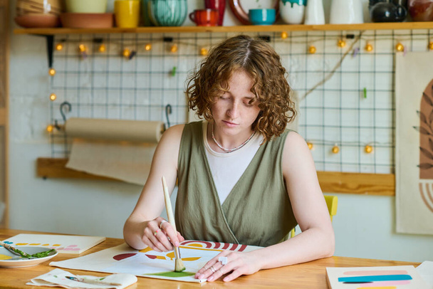 Joven mujer moderna en ropa casual sentada junto a la mesa y trabajando sobre la creación de una nueva pintura abstracta en el taller o estudio - Foto, Imagen