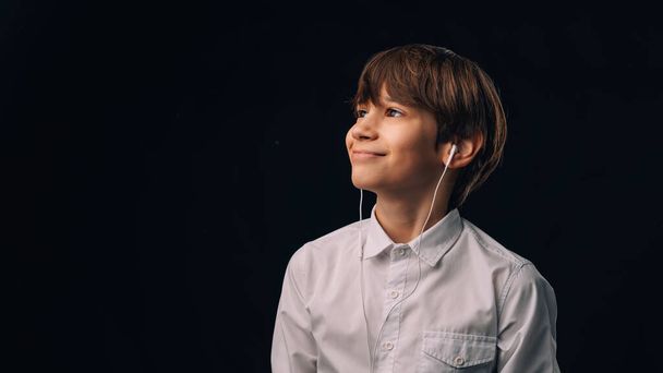 Knappe tiener jongen draagt witte shirt is het luisteren naar de muziek in hoofdtelefoon over zwarte achtergrond. - Foto, afbeelding
