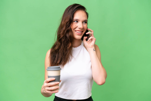 Νεαρή καυκάσια γυναίκα απομονωμένη σε απομονωμένο φόντο κρατώντας καφέ για να τον πάρει και ένα κινητό - Φωτογραφία, εικόνα